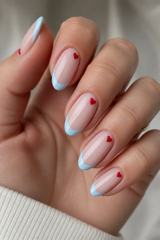 Medium almond nails with a sheer nude base, sky-blue French tips, and a tiny red micro heart tucked at the cuticle corner on each nail.