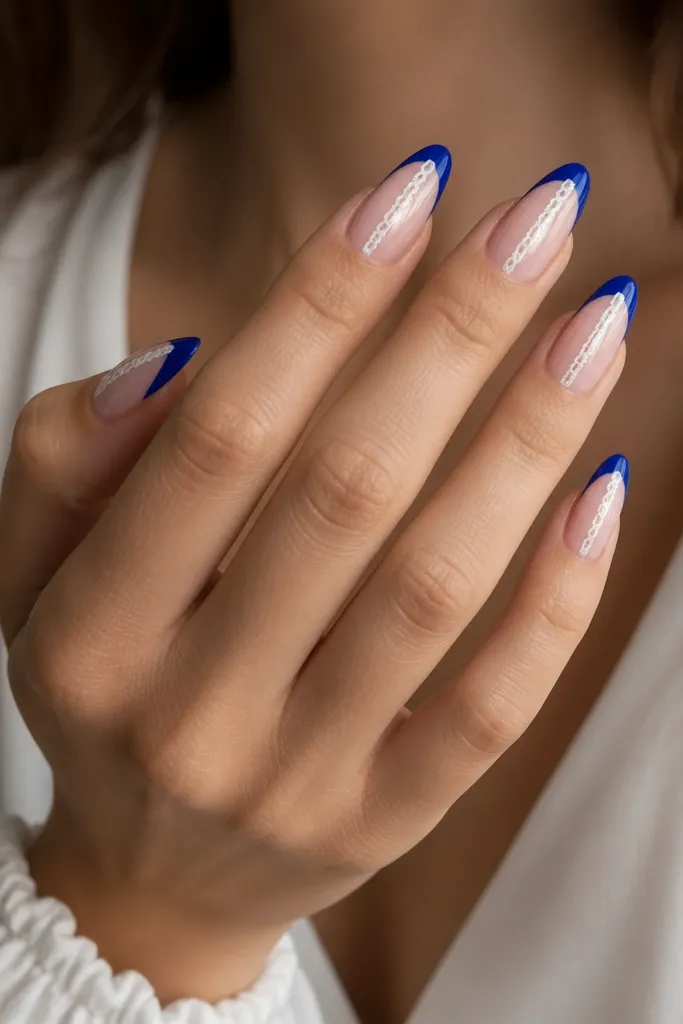 Long-ish almond nails with a sheer nude base, royal-blue French tips, and a single vertical white lace-like stripe running from the tip edge toward the center.