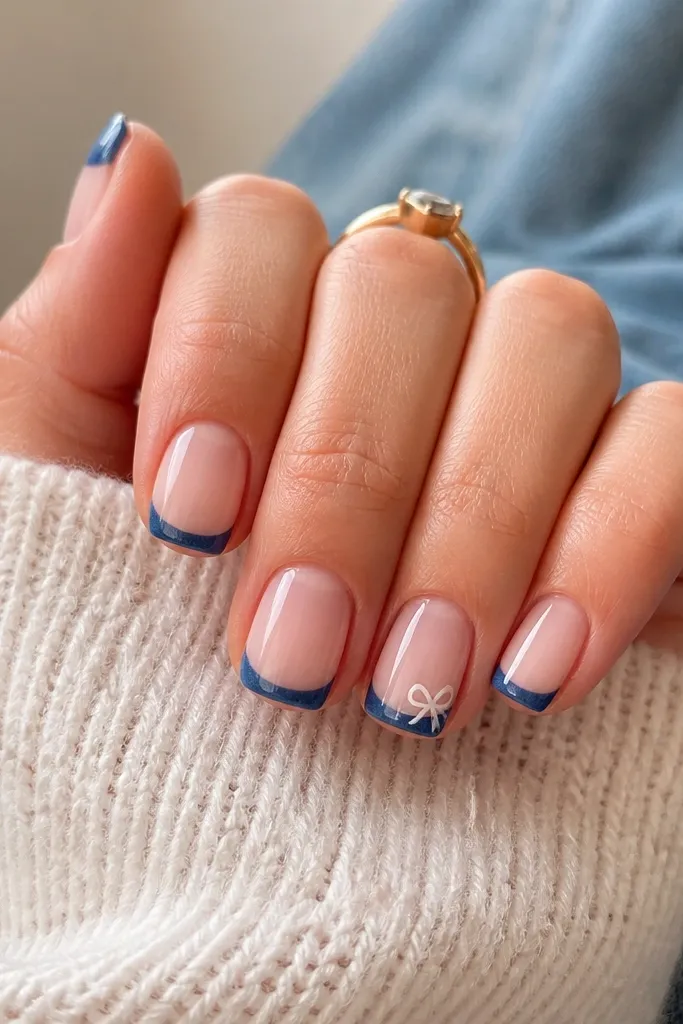 Short squoval nails with denim-blue French tips; on the ring finger, a small white bow sits on the outer side of the tip.