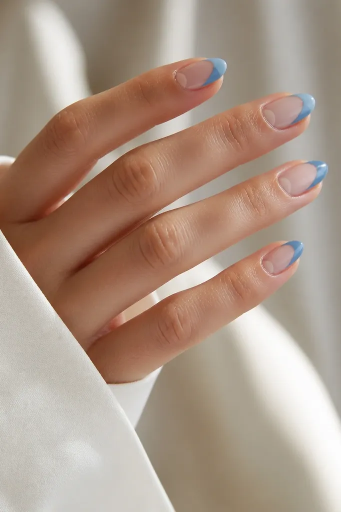 Medium almond nails with a nude base; blue French tips are painted only on one half, leaving a negative-space half-moon near the cuticle.