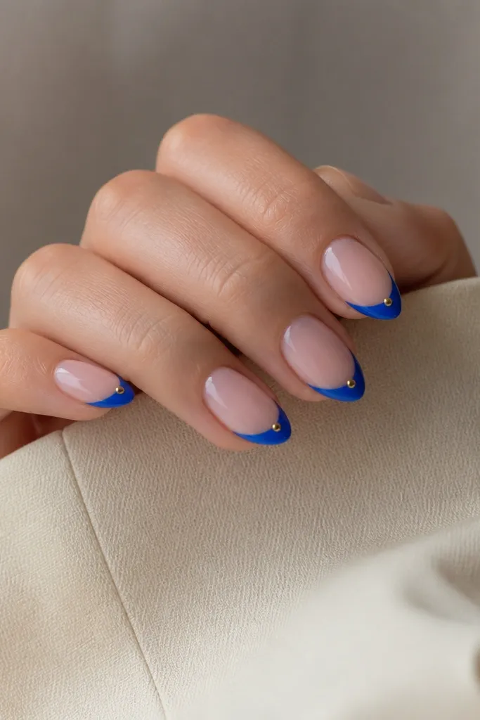 Medium almond nails with bright blue French tips and a single tiny gold stud placed at the outer corner of each tip.