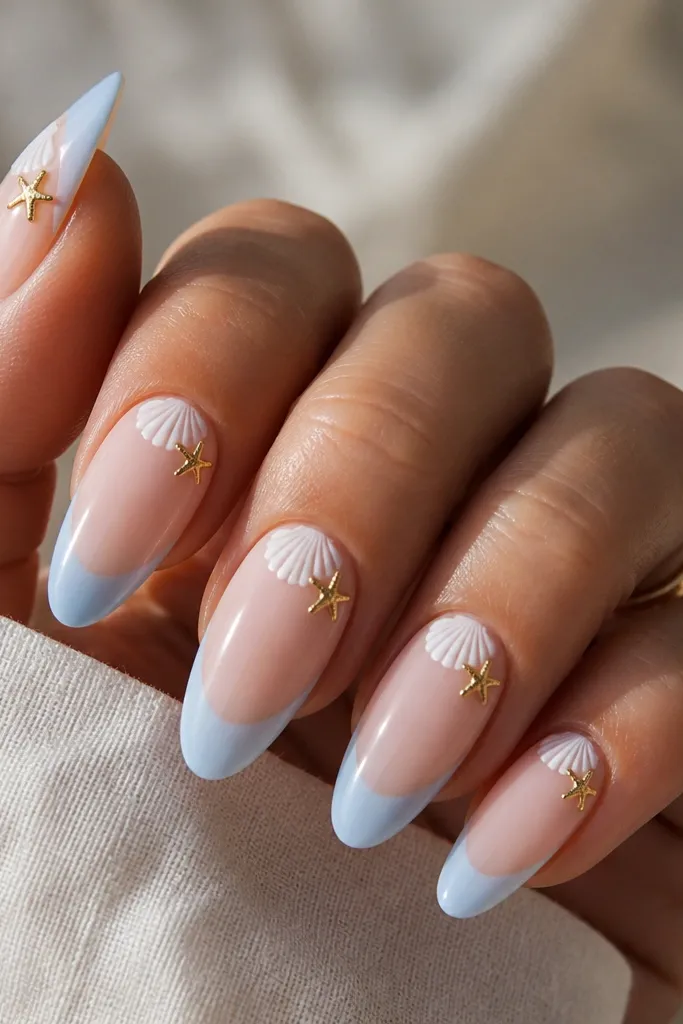 Long almond nails with a soft blue French tip. The base is sheer nude. A small white shell is painted near the cuticle, and tiny gold starfish charms sit at the side of each nail.
