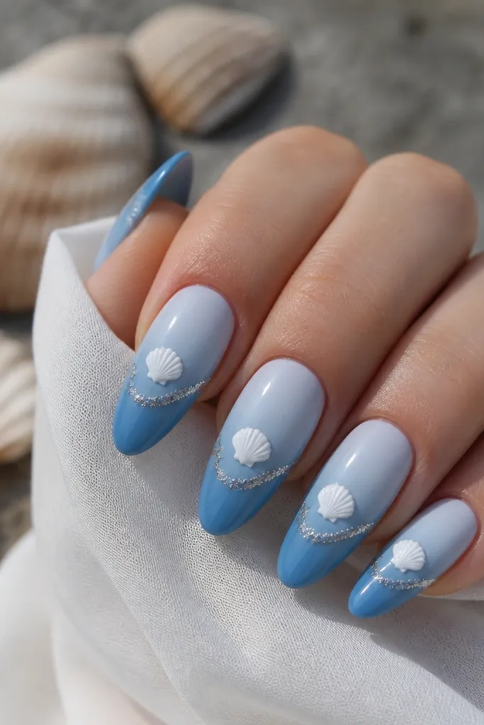 Long almond nails painted a gradient from pale blue at cuticle to deeper blue at tip. A white shell sits centered, and a glittery wave line runs under the shell.