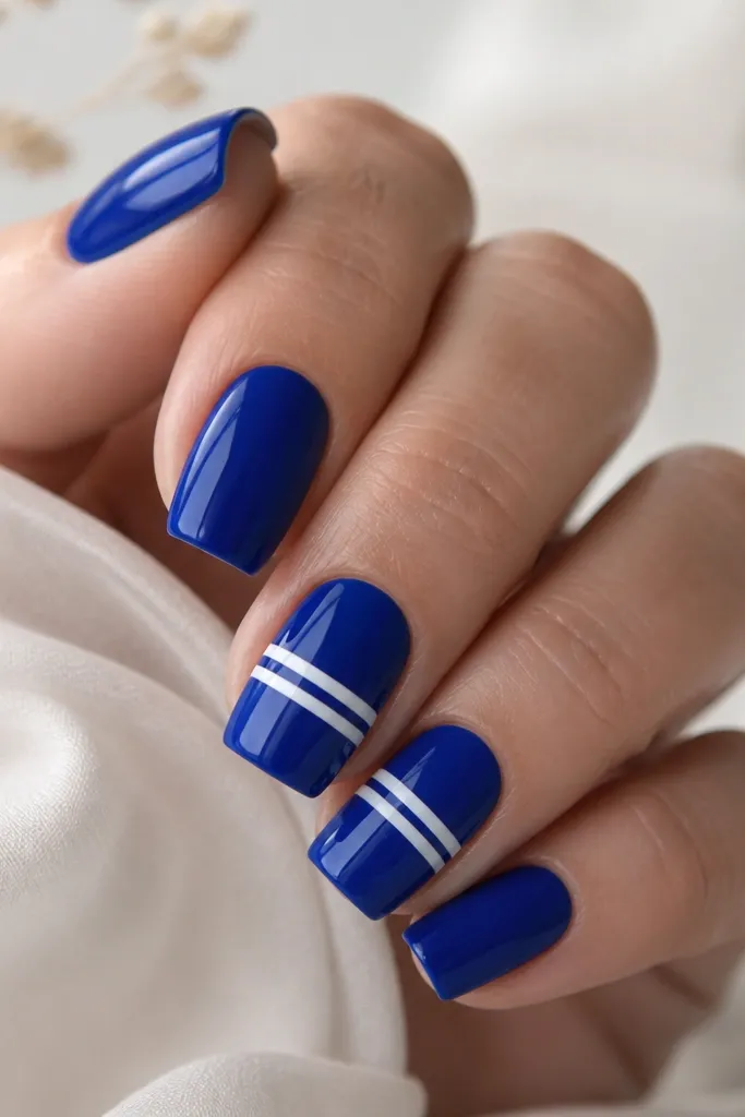 Square nails painted glossy cobalt blue. Two nails have thin white stripes made with nail art tape across the middle, leaving clean gaps. Other nails are solid blue.
