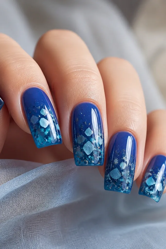 Square nails in deep blue jelly gel, glossy and translucent. Small sea-glass shaped confetti pieces are embedded near the middle and tip. The confetti is light aqua and white with a few silver flecks.