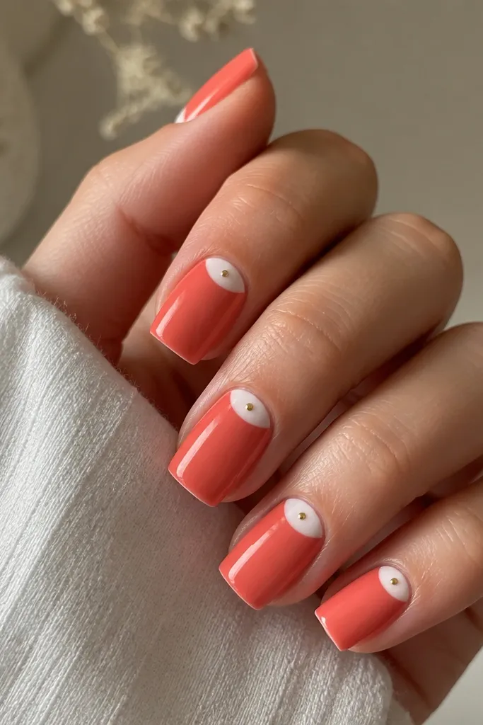 Square nails with a coral solid base. Each nail has a white half-moon/half negative-space shape on one side of the nail near the cuticle. A single tiny gold dot sits near where the shapes meet.