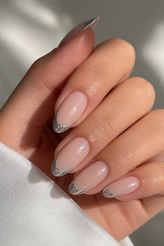 Almond nails with a sheer nude base. The French arc is silver chrome. At each outer corner of the smile, there are two tiny clear crystals set like a V shape.