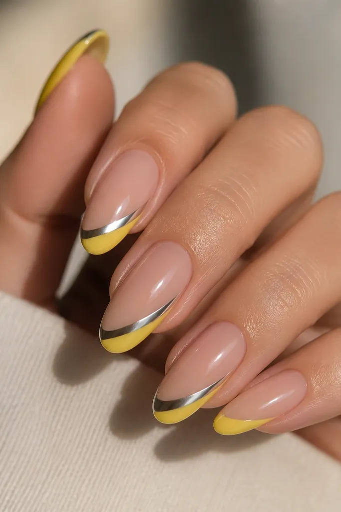 Long oval nails. Yellow French tips are smooth and glossy. A chrome half-glaze sits diagonally across each tip, leaving the other side plain yellow. Silver chrome, nude base.