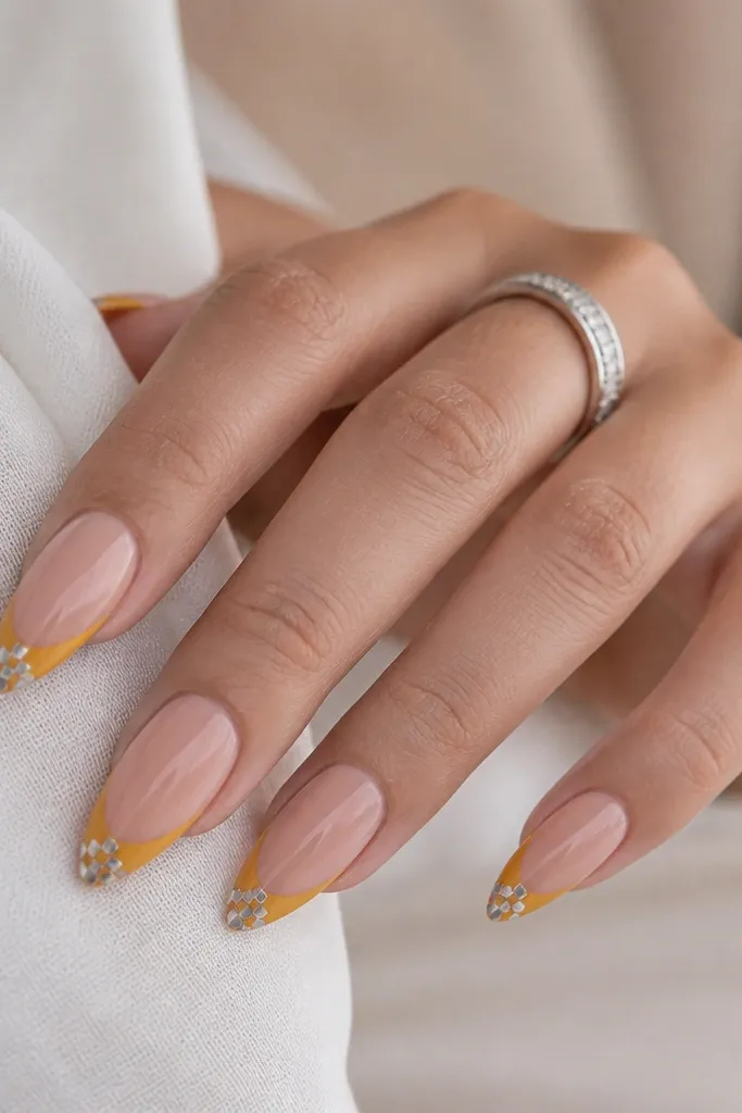 Stiletto or long almond nails. Mustard yellow French tips. The free edge has a tiny checker pattern in silver chrome, like miniature squares. Nude base, glossy.