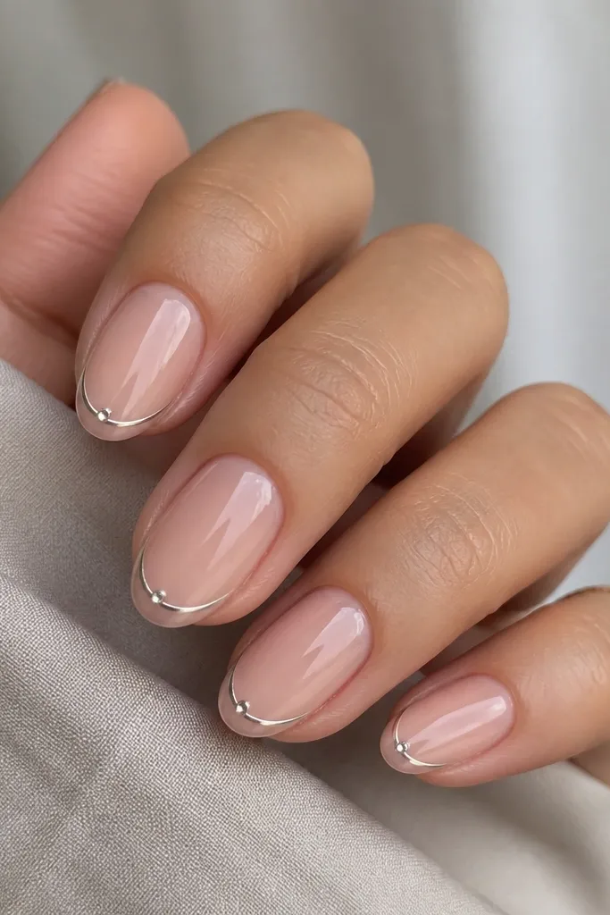 Short almond nails with a blush-pink base. The French tip is a thin chrome line in mirror silver. At the center of the smile line (where the French curve begins), there's a single tiny chrome crystal dot that sparkles.