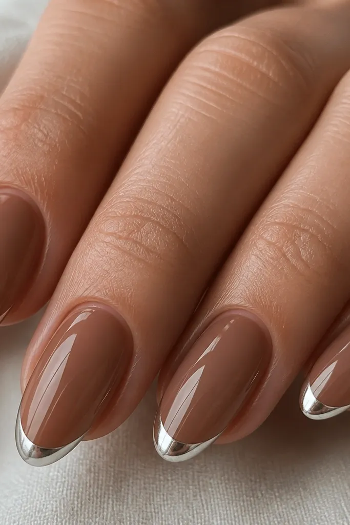 Close-up of medium almond nails. The base is a sheer mocha-brown nude with a smooth jelly finish. The French tips are very narrow, about a thin curved band across each nail, in bright mirror silver chrome with razor-sharp edges. The cuticles look clean, no flooding onto skin.
