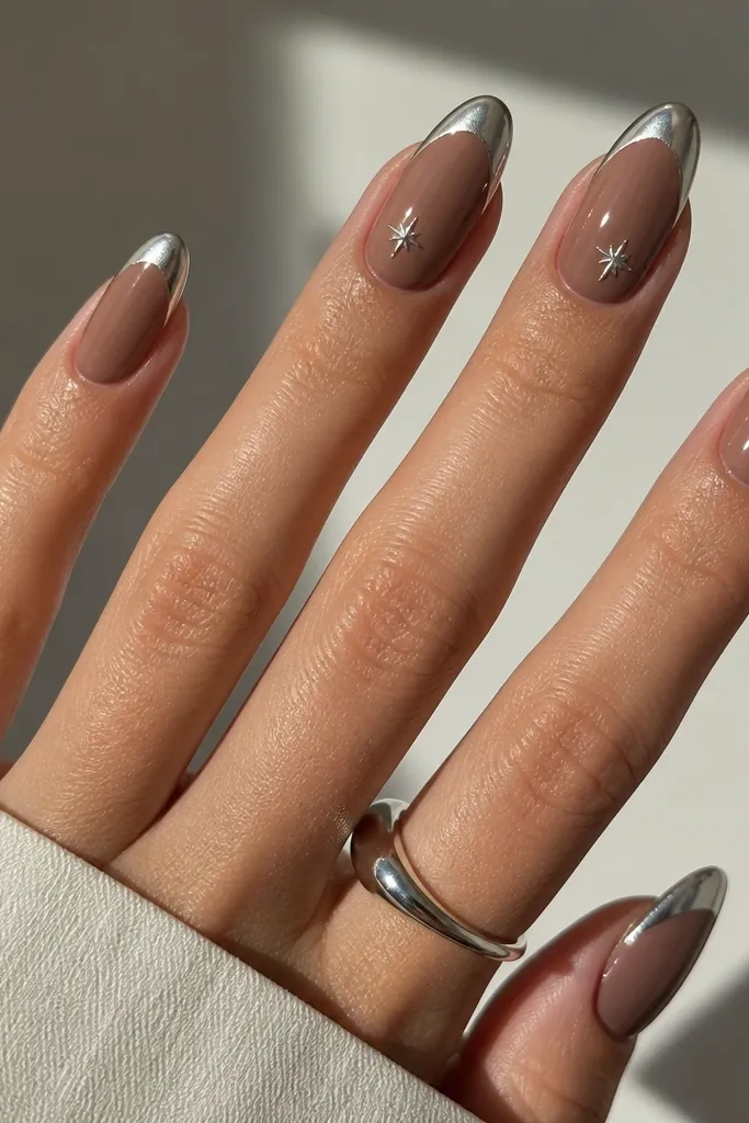 Almond nails with a cocoa nude base. The French tips are mirror chrome silver, medium width. On two nails, there are tiny star accents at the side near the mid-nail, one per side, in silver chrome. The rest of the nails are clean chrome French only.