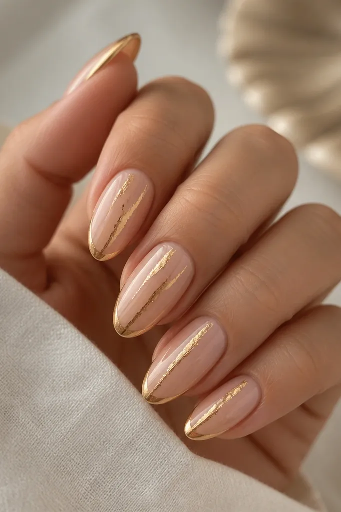 Medium almond nails; nude base with two slim vertical gold foil streaks leading toward the French tip; gold mirror French line at the free edge; glossy finish.