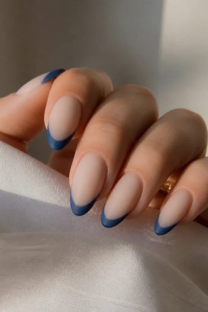 Almond nails with a milky nude base, deep blue French tips, and a matte top coat with a soft, velvety look.