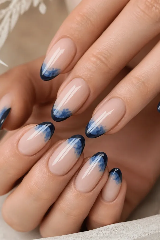 Nude base nails with dark navy French tips and a light blue watercolor splash at the center of the tip, airy and artistic, glossy finish.