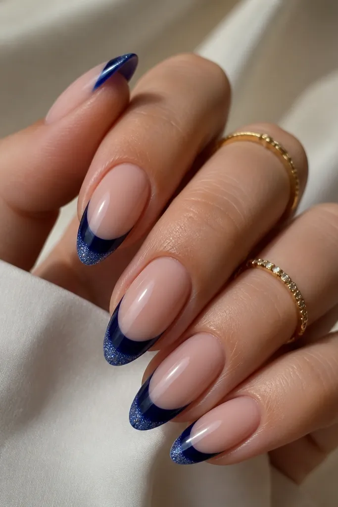 Almond nails with navy French tips that fade into a fine sparkly blue at the extreme tip edge, glossy finish.