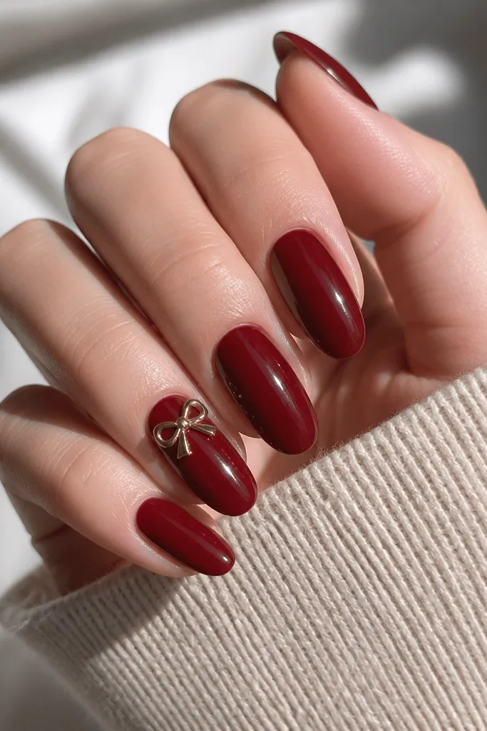 Glossy dark cherry red nails with one accent nail featuring a small 3D bow charm near the cuticle. The charm is metallic with a soft highlight, and the rest of the nails are plain.