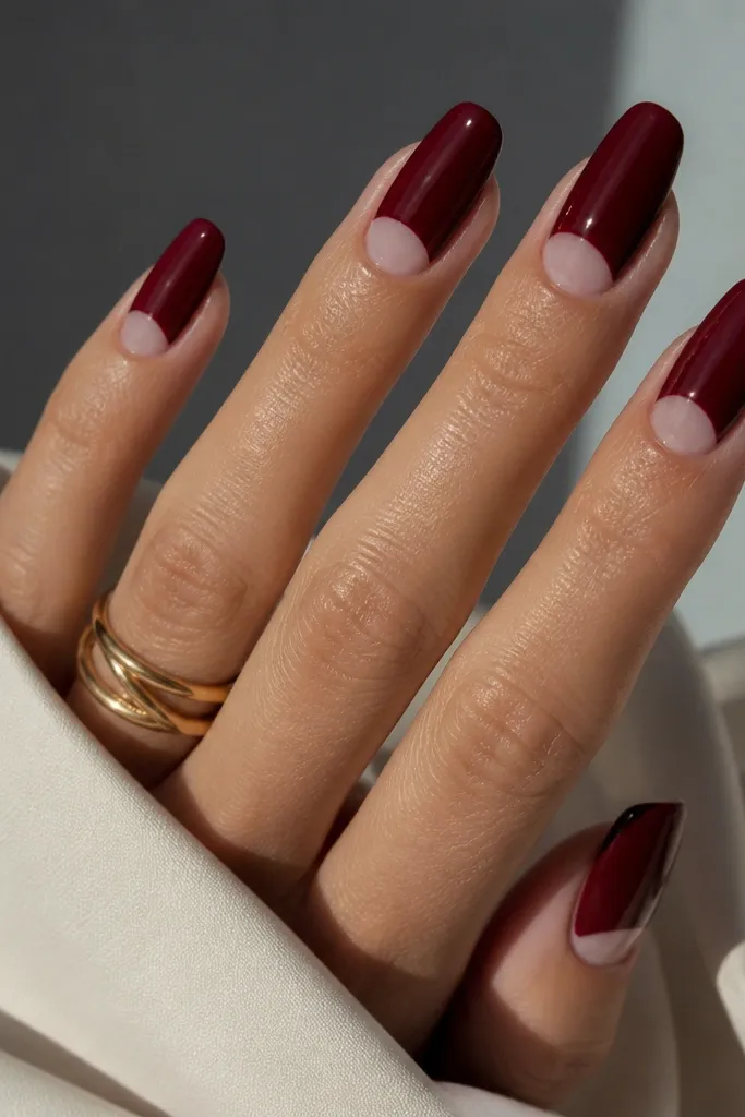 Dark cherry red acrylic nails with a negative space half moon near the cuticle. The bare nail area is visible as a clean crescent, and the red starts below it. Glossy finish.