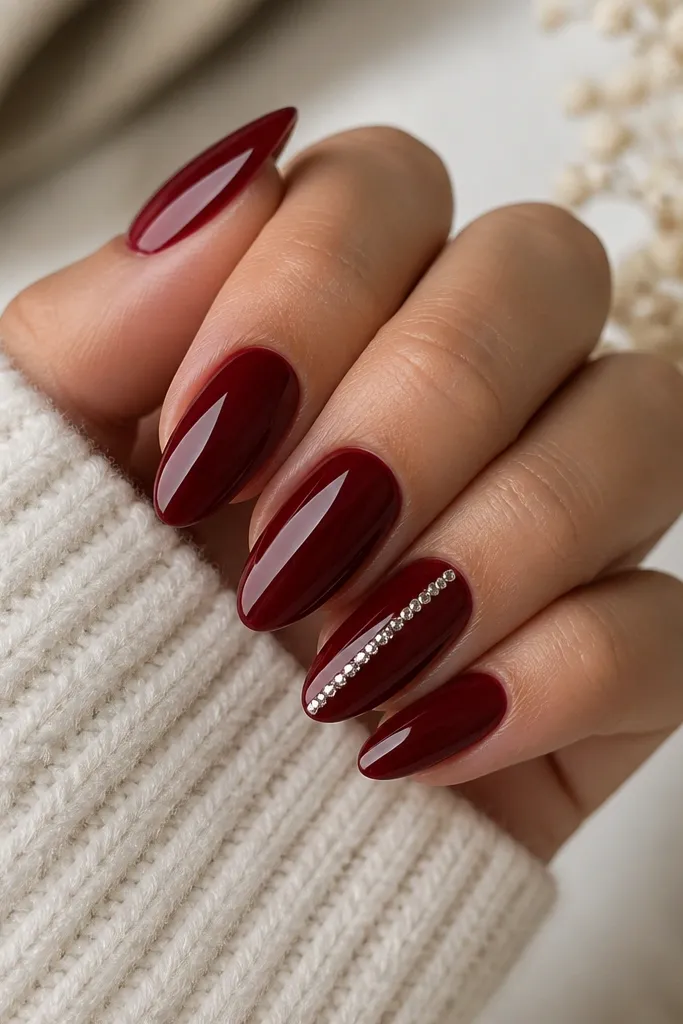 Most nails are solid dark cherry red glossy; one accent nail has a vertical column of tiny rhinestones from near the cuticle toward the tip, almond shape.