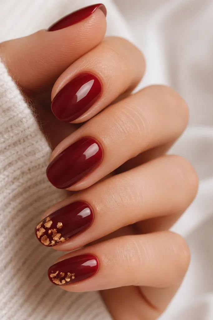 Solid dark cherry red nails with one or two nails featuring gold leaf shatter pieces across the tip area, glossy.