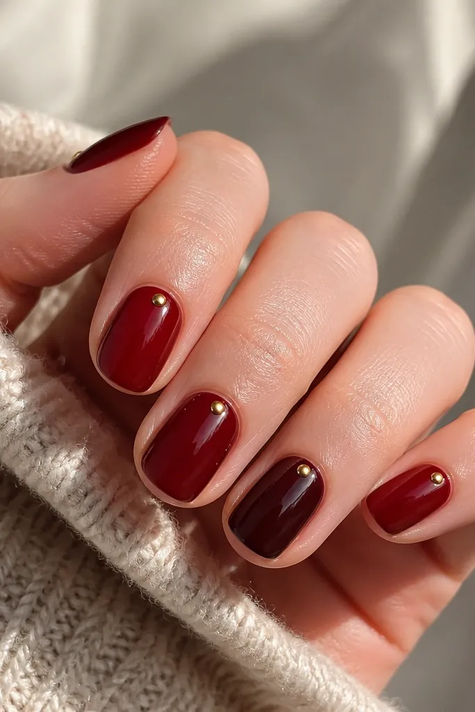 Four nails in dark cherry red and one in oxblood maroon, each with tiny gold dot accents near the cuticle, short almond/squoval.