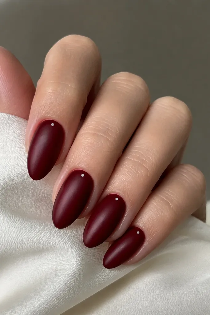 Dark cherry red nails with a satin-sheen finish and tiny white dots placed near the sidewalls, almond shape.