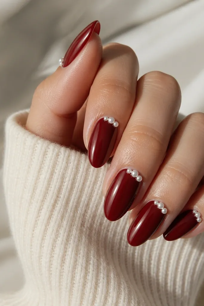 Dark cherry red nails with 2-3 small pearl beads clustered along one side of the cuticle on each nail, glossy, almond shape.