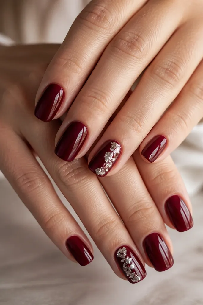 Glossy dark cherry red nails with one accent nail per hand. The accent nail has small silver foil flakes clustered near the outer edge, like crushed metal. The rest of the nails are solid cherry.