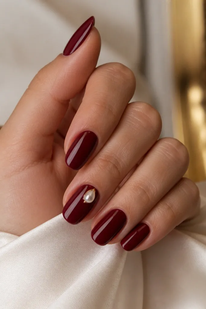 Glossy dark cherry red nails with one accent nail that has a small white pearl teardrop gem set near the outer sidewall close to the cuticle. The gem catches light and looks dimensional.