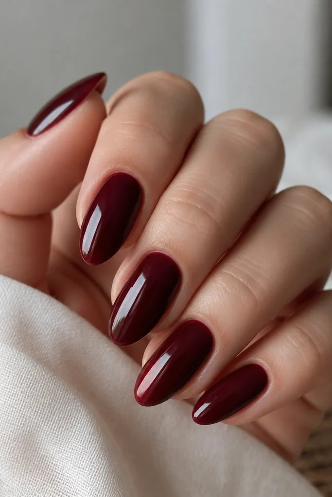 A close-up of medium-length almond nails painted a deep dark cherry red with a high-gloss mirror shine. The cuticles are clean, and each nail has a smooth, even color with no glitter or art.