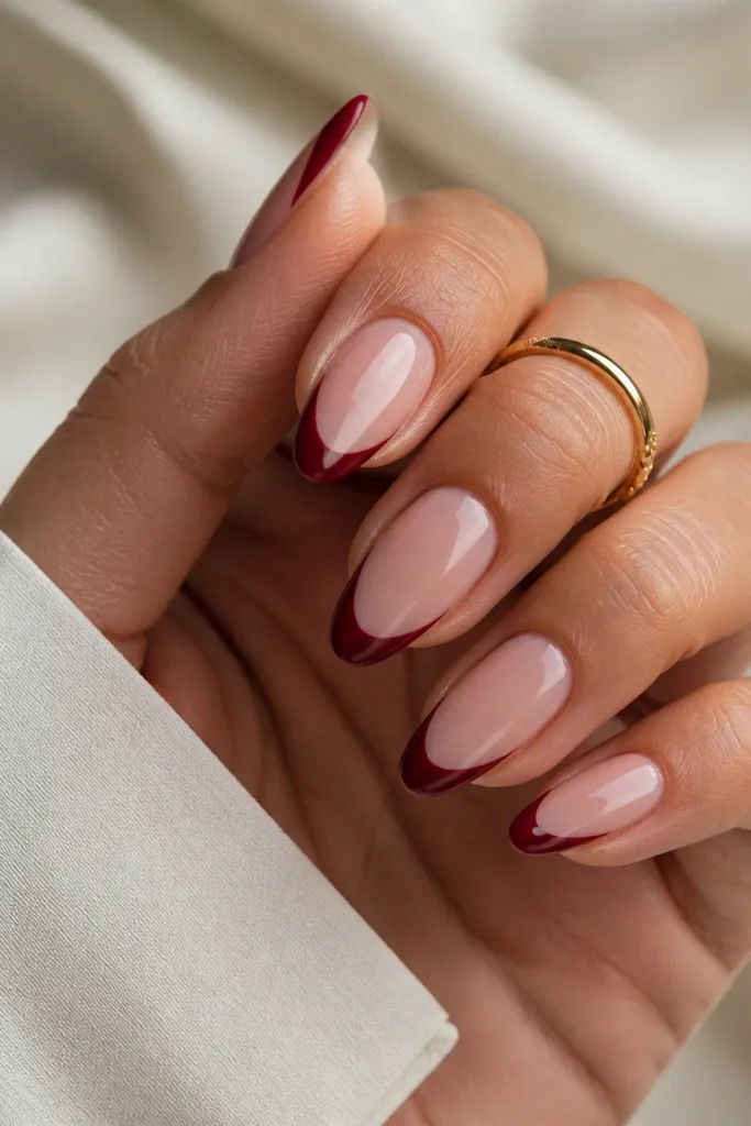 Almond nails with a nude pink base and crisp dark cherry red French tips. The tips form a sharp arc at the free edge with clean negative space at the sidewalls.