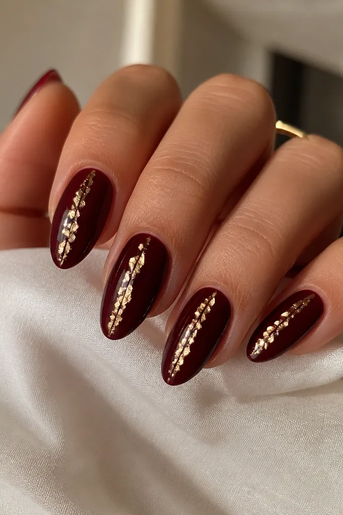 Dark cherry red almond nails with thin, irregular gold foil lines running vertically near the center of each nail. The gold catches light like shards without covering the whole nail.
