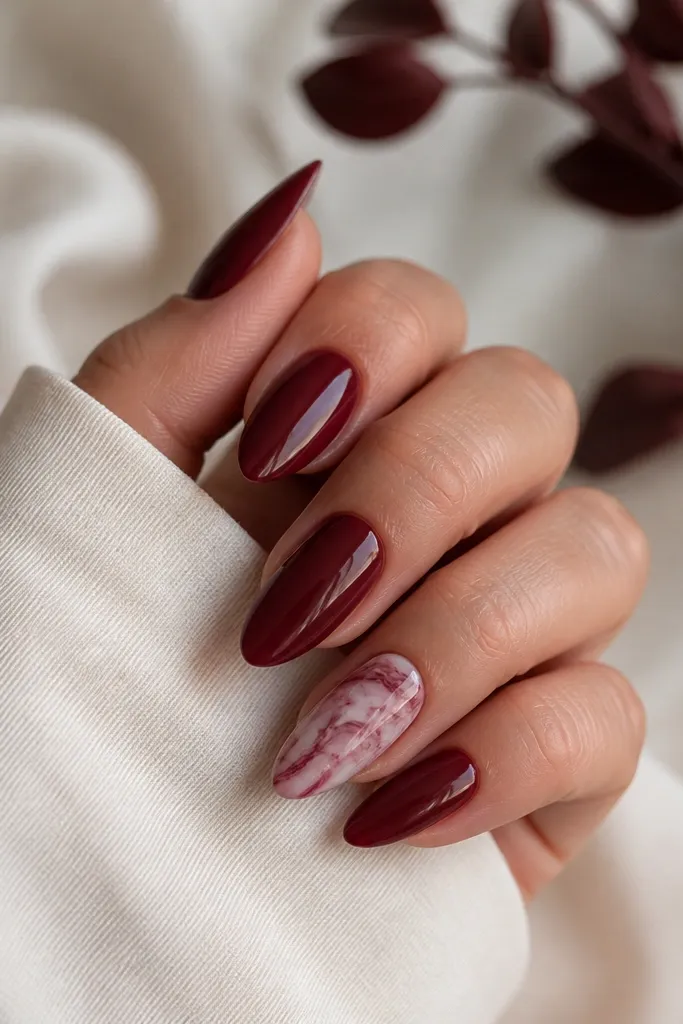 Almond nails in dark cherry red with one accent nail featuring white and deep burgundy marble swirls. The marble lines look thin and vein-like, not chunky.