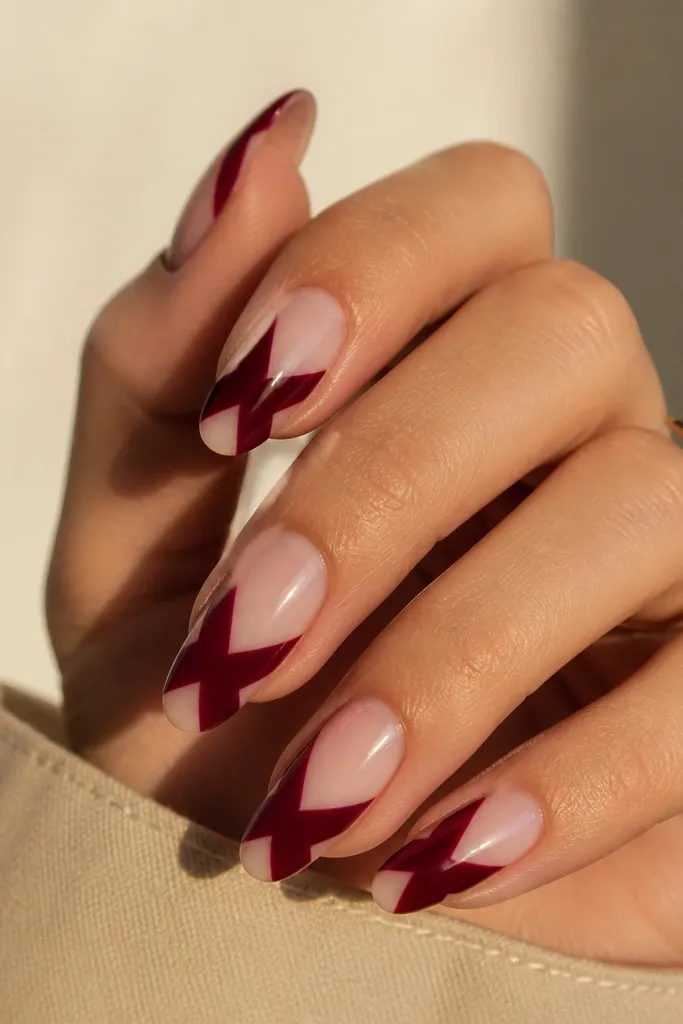 Almond nails with dark cherry red forming diagonal crisscross shapes, leaving small negative space triangles near the base and sidewalls. The rest of the nail is bare or sheer nude.