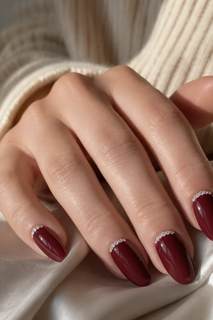 Dark cherry red almond nails with a small half-moon crescent of tiny rhinestones at the base near the cuticle. The rest of each nail is glossy plain cherry.
