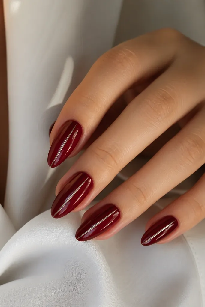Almond nails painted dark cherry red with one thin vertical silver stripe running down the center of each nail. The stripe is crisp and reflective, and the rest is glossy plain cherry.