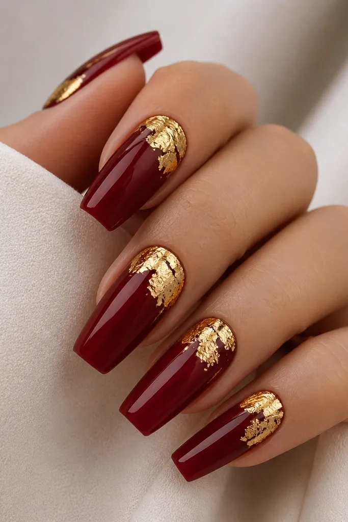 Glossy dark cherry red coffin nails with irregular gold leaf pieces placed near the cuticle line on each nail, leaving some bare cherry visible around the leaf.