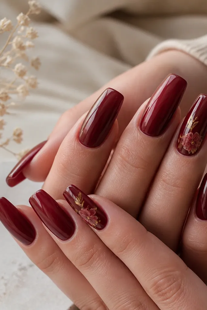 Coffin nails mostly glossy dark cherry red; one or two nails have a small hand-painted floral design in lighter red and gold near the tip.