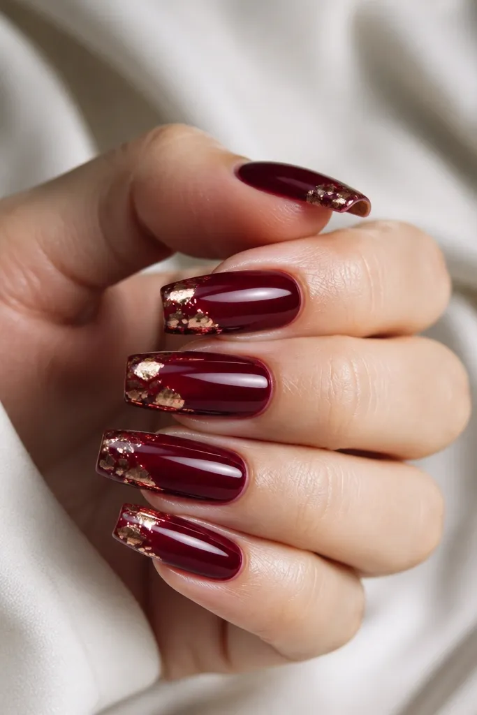 Coffin nails glossy dark cherry red with a strip of metallic foil rubbed at the tip, showing irregular gold and rose-toned foil fragments.