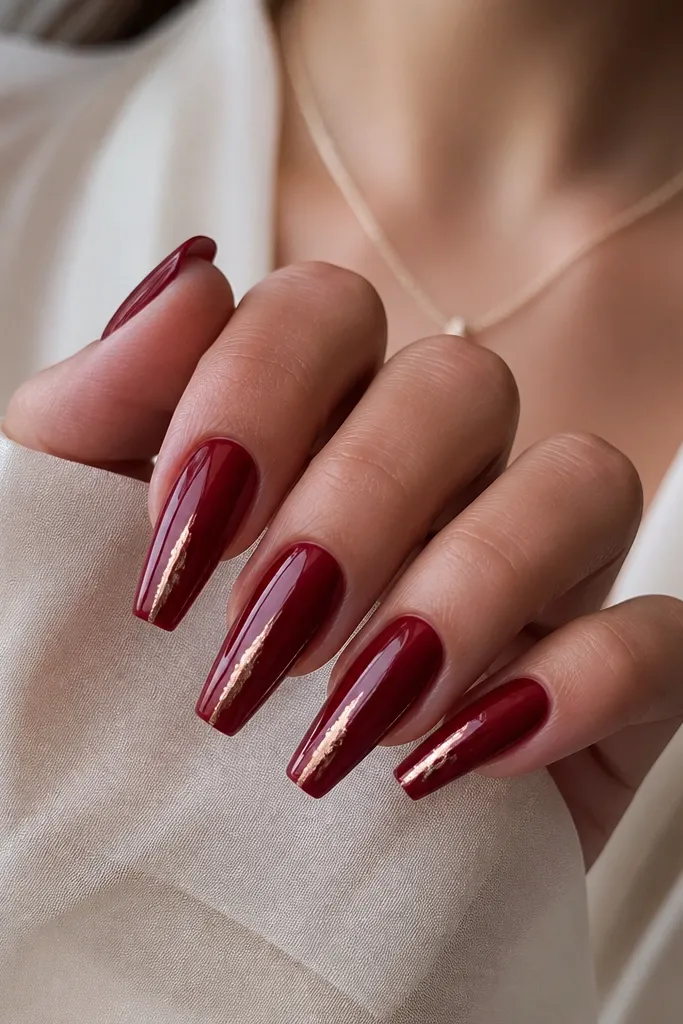 Glossy dark cherry red coffin nails with thin rose-gold foil lines running vertically toward the tip, like burnished streaks.