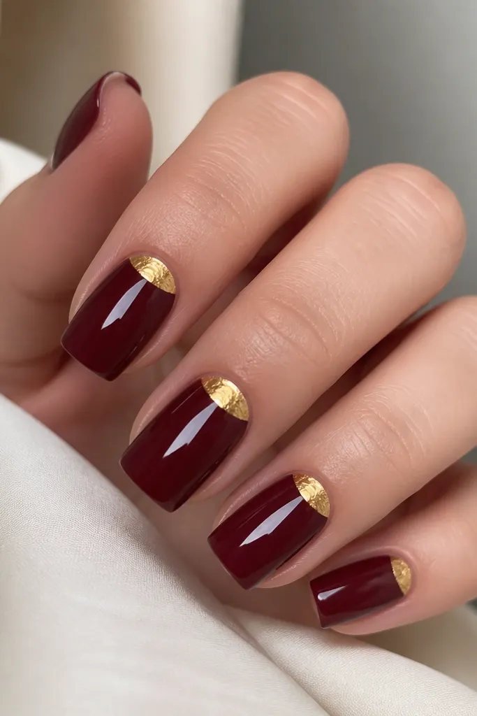 Dark cherry red square nails with a metallic gold half-moon at the cuticle area on each nail, glossy red base, gold foil that looks slightly textured, clean cuticle line.