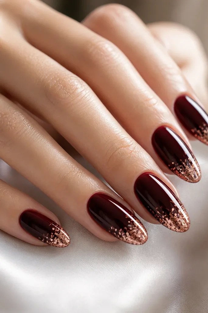 Dark cherry almond nails with a French tip covered in rose gold foil flakes. The foil is concentrated at the tip edge and thins toward the smile line, with a glossy finish over everything.