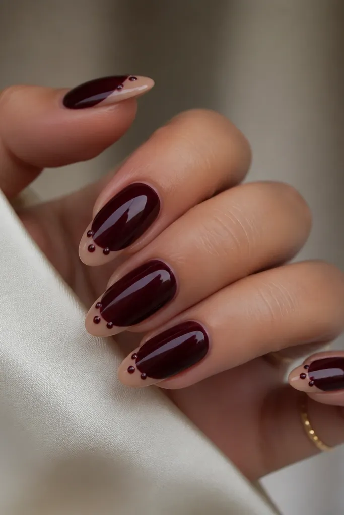 Almond nails in dark cherry red with a nude French tip. Along the smile line, three tiny burgundy studs sit like a row, centered on each nail, with high shine.