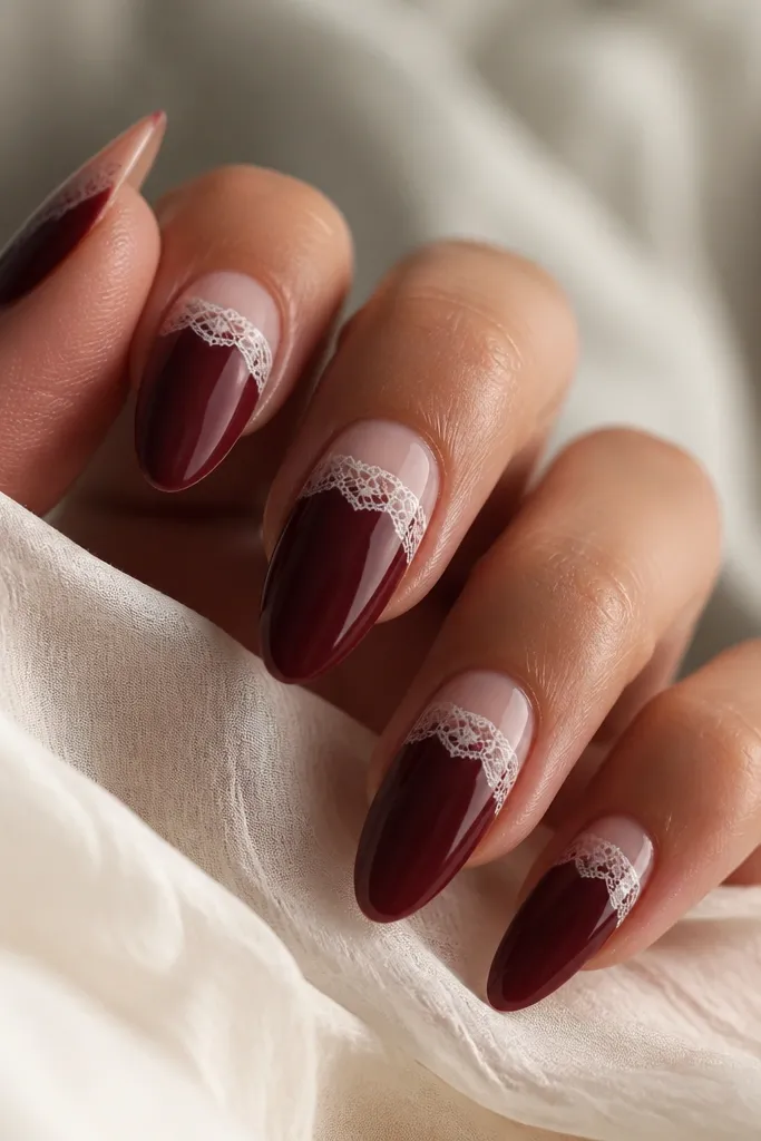 Almond nails with dark cherry red base. The French tip is nude, and a small piece of white lace sits right at the smile line, with lace pattern visible and sealed under glossy gel.