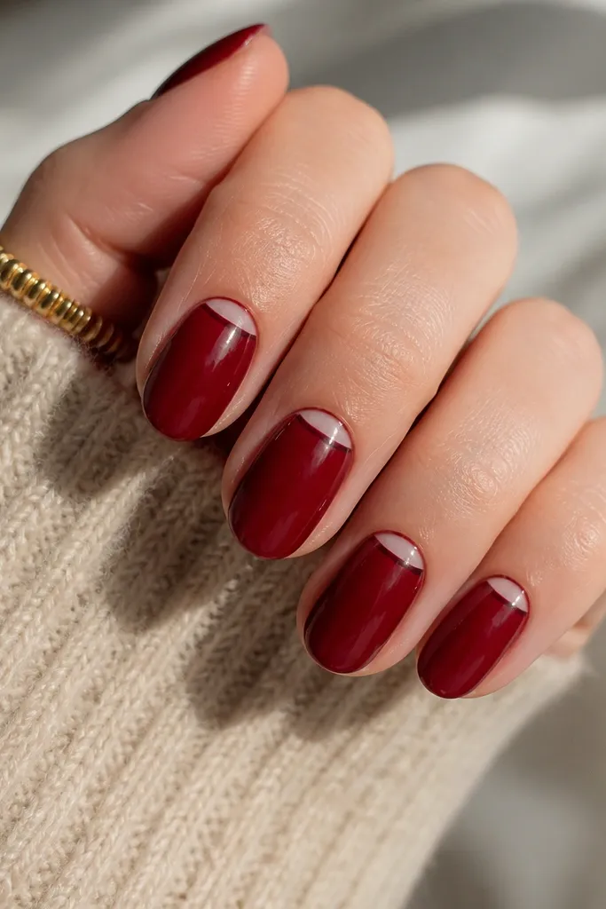 The nails have a deep cherry red full base in glossy finish. Instead of a white tip, there's a reverse French at the cuticle: a curved, slightly darker wine-red arc outlined cleanly, leaving a small clear crescent near the cuticle. The rest of the nail is smooth and shiny.