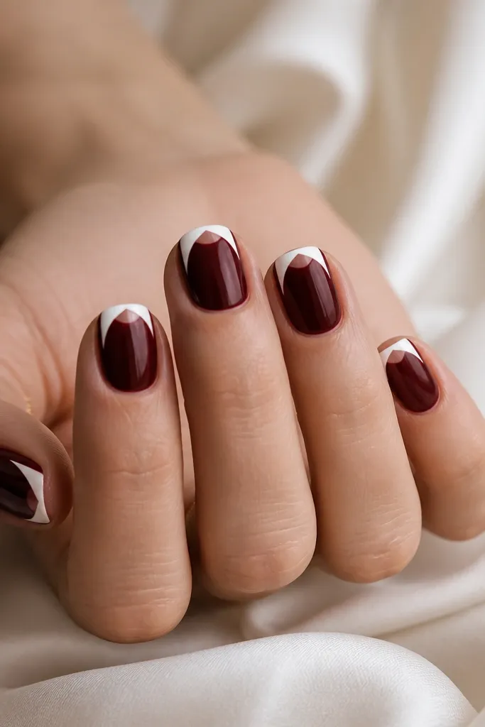 Short, square-rounded nails. The base is dark cherry red glossy. The French tip is a clean white outline that forms a V shape, but the center of the tip area stays clear, leaving a half-moon negative space that looks like the nail is cut out.