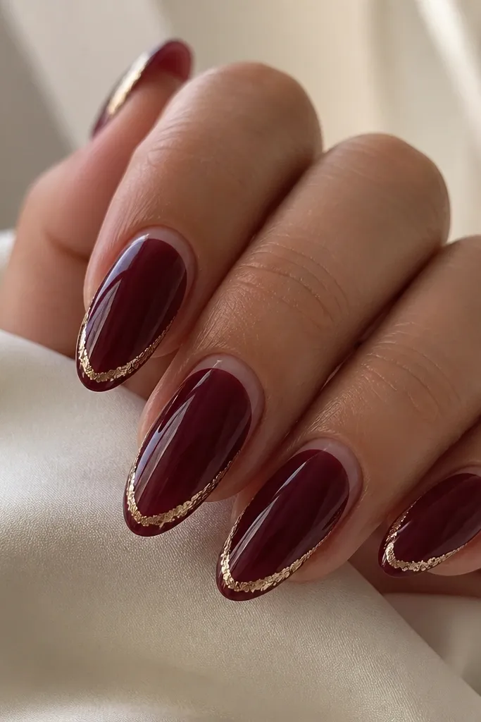 Medium-length almond nails painted glossy dark cherry. The French tip is a thin, clear-to-cherry transition with a gold foil strip running along the outer curve of the tip, like a metallic frame. The gold pieces are irregular foil fragments but arranged in a clean line.