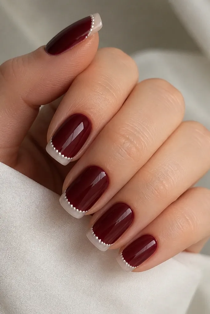 Glossy dark cherry nails with a French tip band in clear-to-white gradient. Along the outer edge of the French tip, there are tiny pearl dots spaced evenly like a beaded bracelet. The dots are small, off-white, and sit in a straight line.