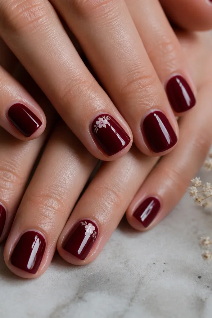 Short nails with dark cherry red base; a tiny white floral stamp sits near the cuticle on two nails; glossy.