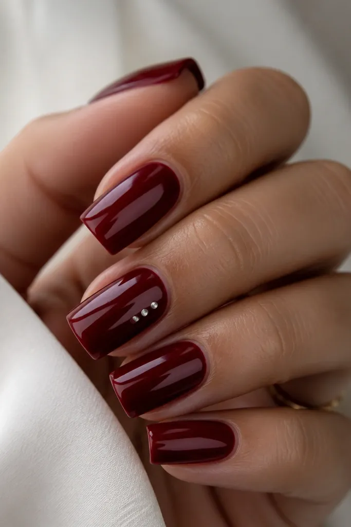 Square nails in dark cherry red with a line of three tiny white dots on the middle finger angled diagonally, glossy top coat.